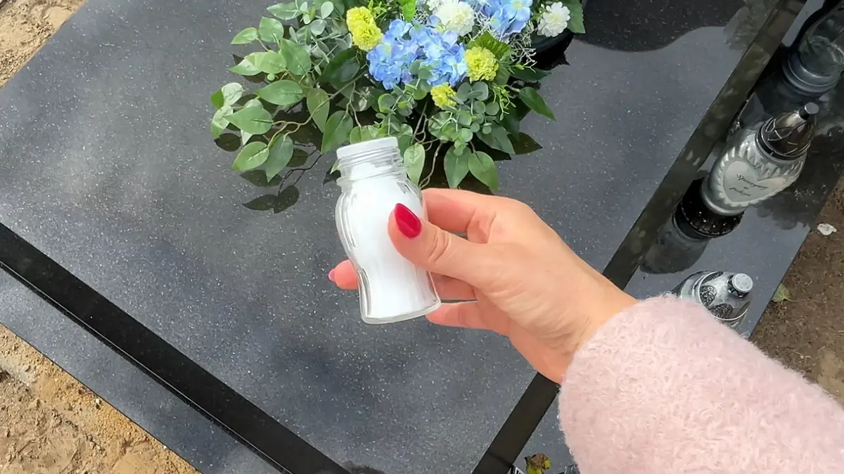 Dłoń trzymająca solniczkę z solą kuchenną nad czarną płytą pomnika cmentarnego.